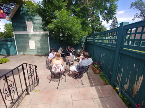 A group of people sitting around a table in a sunny outdoor patio, surrounded by greenery and a wooden fence.