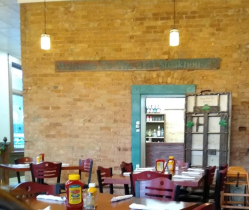 Interior of a rustic steakhouse with wooden tables, chairs, and a brick wall featuring a welcome sign.