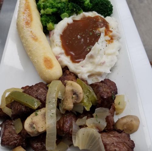 A plate featuring sautéed beef with onions and mushrooms, mashed potatoes with gravy, steamed broccoli, and a breadstick.