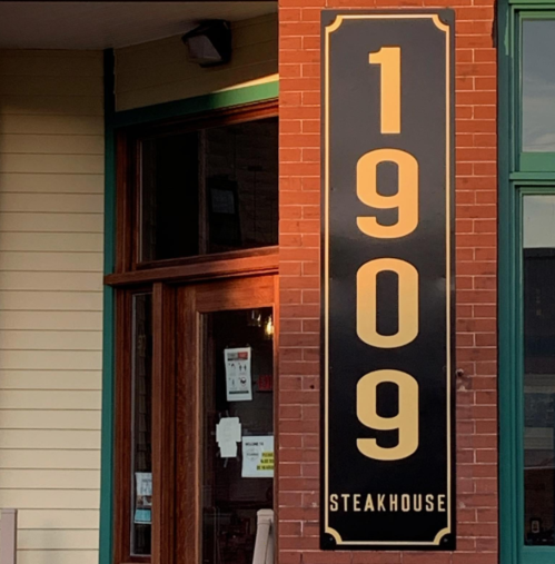 Sign for "1909 Steakhouse" displayed on a brick wall, featuring bold gold lettering.