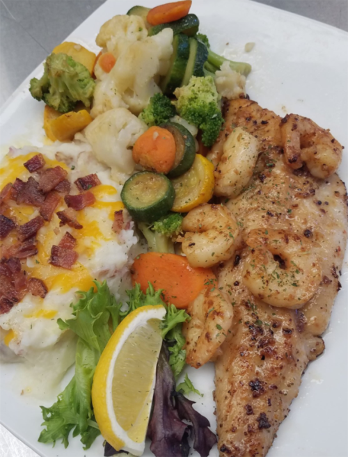 A plate featuring grilled fish, shrimp, mixed vegetables, cheesy rice with bacon, and a lemon wedge.