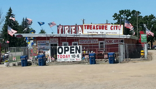 A colorful storefront of "Treasure City" with flags, a picnic area, and an open sign displaying hours.