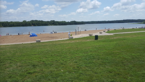 A sandy beach by a lake with people enjoying the water, green grass in the foreground, and a cloudy sky above.