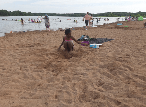 A child sits in the sand at a beach, with people swimming and relaxing in the background.
