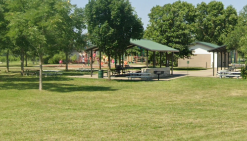 A sunny park scene featuring picnic areas, trees, and a playground in the background.