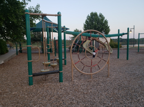 Playground equipment with climbing structures and a spider web climber, surrounded by gravel and trees at dusk.