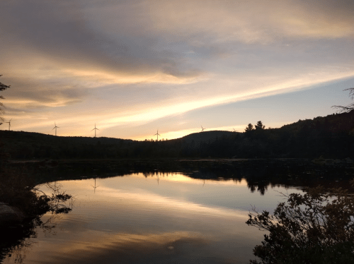 A serene sunset over a lake, with silhouettes of wind turbines on the distant hills and colorful clouds in the sky.