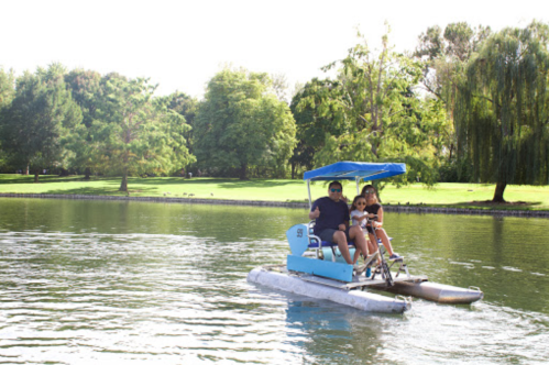 A man and a girl ride a blue pedal boat on a calm lake surrounded by trees and greenery.