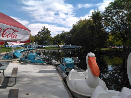 A serene lakeside scene with paddle boats, a large swan-shaped boat, and trees under a partly cloudy sky.