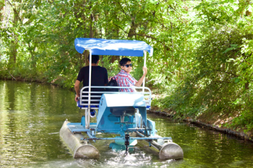 Two people on a blue pedal boat in a lush, green canal, enjoying a sunny day.