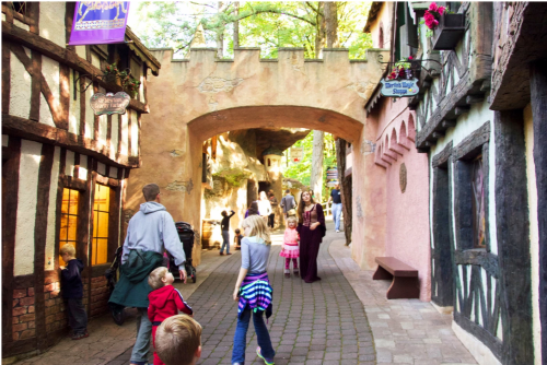 A charming village street with people walking, featuring medieval-style buildings and colorful decorations.