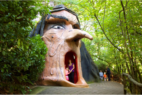 A whimsical giant witch's head sculpture with an open mouth, surrounded by trees, as children and adults explore nearby.