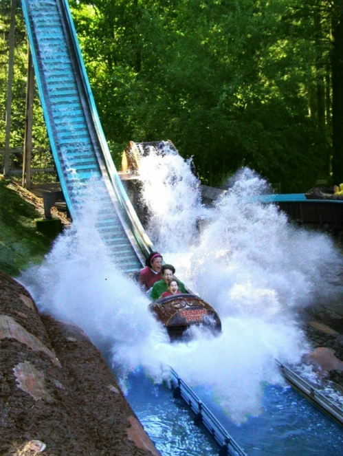 A log flume ride splashes down, sending water high as two riders enjoy the thrill in a lush, green setting.