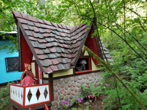 A whimsical miniature house with a stone facade, red accents, and a doll on the porch, surrounded by lush greenery.