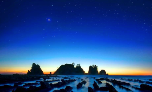 Silhouette of rocky coastline under a starry sky at dusk, with calm ocean waves reflecting the twilight colors.