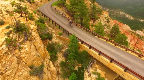 A cyclist rides along a winding road on a cliffside, surrounded by trees and rocky terrain. Scenic mountain views in the background.