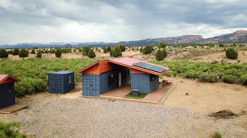 A modern cabin with solar panels, surrounded by desert landscape and mountains in the background.