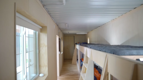 Interior of a compact living space featuring a lofted bed, wooden flooring, and large windows for natural light.