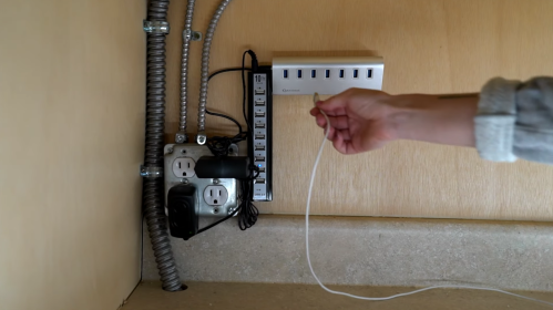 A hand holds a white cable near a power strip and electrical outlets mounted on a wall.