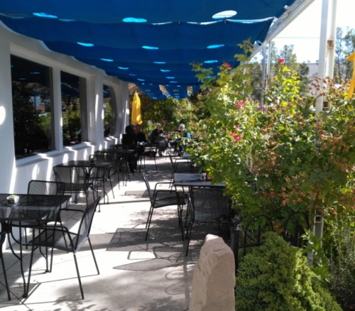 Outdoor dining area with black metal tables and chairs, shaded by a blue canopy and surrounded by greenery and flowers.