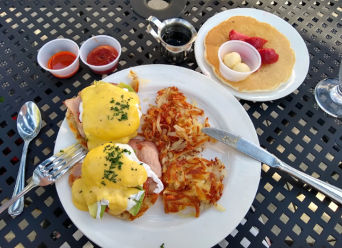A plate of eggs Benedict with hash browns, accompanied by syrup and a small dessert of pancakes with fruit.