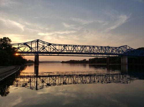 A blue bridge spans a river at sunset, reflecting in the calm water below, surrounded by trees and a serene sky.