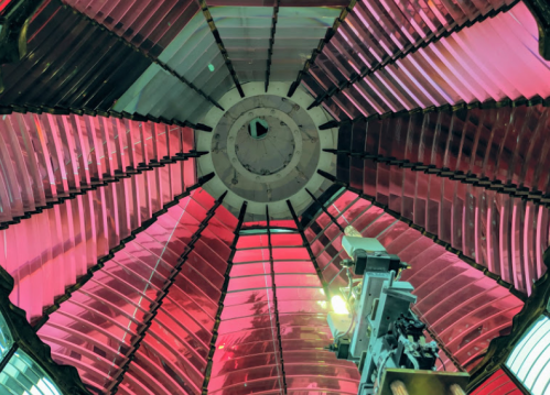 View from below a colorful lighthouse lens, showcasing intricate glass panels in shades of red and pink.