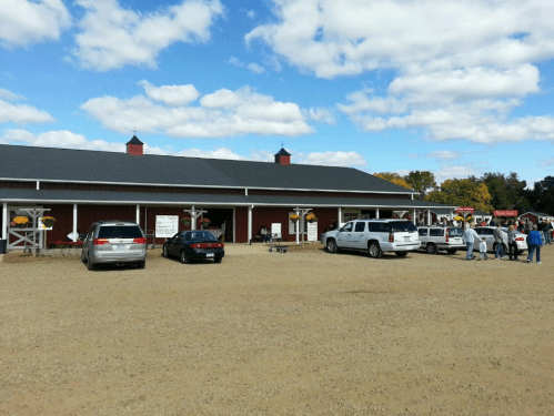 A red barn-style building with cars parked outside, under a blue sky with scattered clouds. People are gathered nearby.