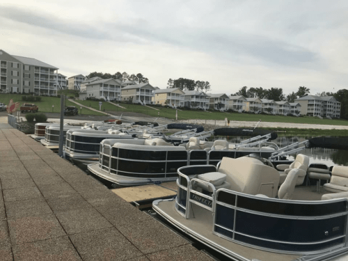 A row of pontoon boats docked at a marina, with waterfront buildings in the background under a cloudy sky.