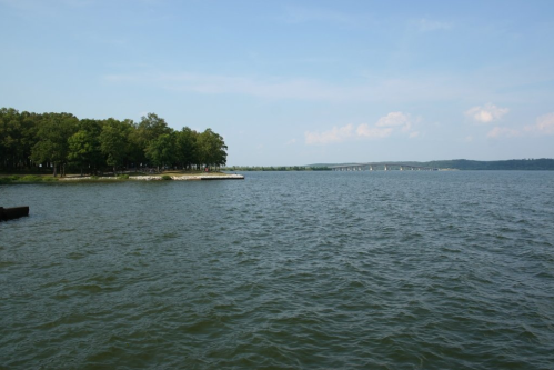 A serene lake view with green trees on the shore and a distant bridge under a clear blue sky.