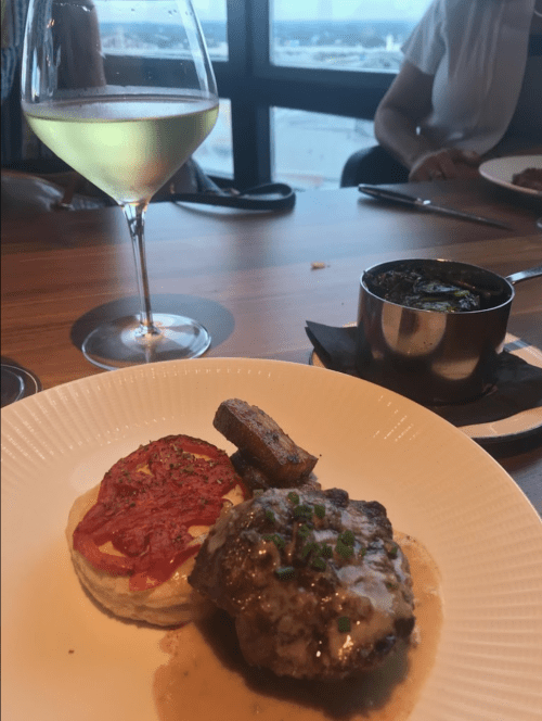 A plate featuring a meat dish with sauce, a slice of tomato, plantains, and a side of greens, accompanied by a glass of white wine.