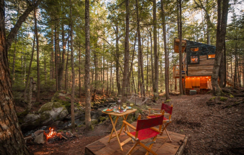 A cozy cabin in a forest, with a wooden deck, chairs, and a small fire pit beside a stream.