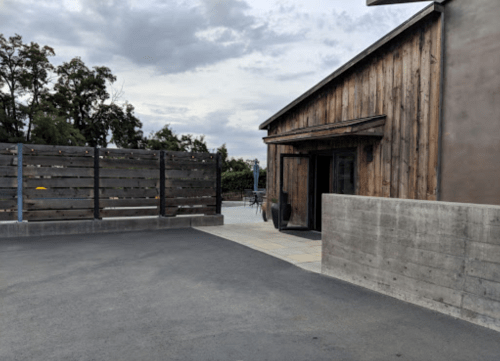 A rustic wooden building with a concrete wall and a paved area, surrounded by trees and cloudy skies.