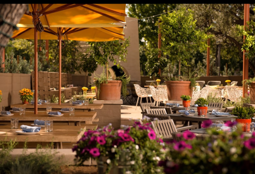 Outdoor dining area with wooden tables, colorful flowers, and yellow umbrellas in a lush garden setting.