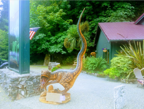 A wooden dinosaur sculpture stands outside a building, surrounded by greenery and an American flag in the background.