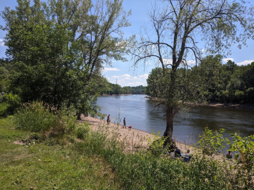 A serene river scene with sandy banks, lush greenery, and people enjoying the outdoors on a sunny day.