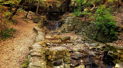 A serene forest scene featuring a stone structure with a small waterfall, surrounded by autumn foliage and rocky terrain.