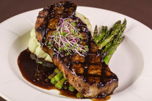 A grilled steak topped with microgreens, served on mashed potatoes and asparagus, drizzled with a rich sauce.