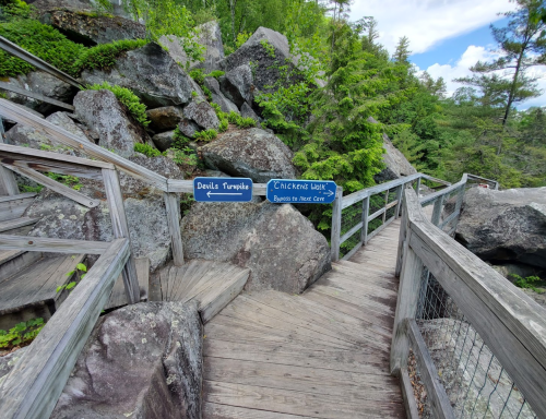 Wooden walkway with signs for "Devil's Turnpike" and "Chicken Walk" surrounded by large rocks and greenery.