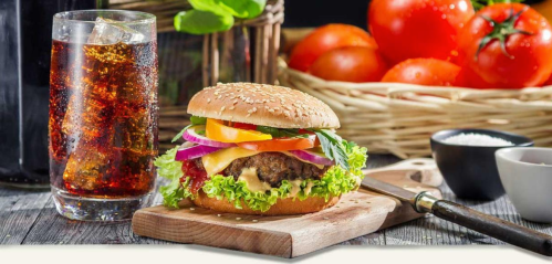 A delicious burger with fresh toppings on a wooden board, accompanied by a glass of soda and a basket of tomatoes.