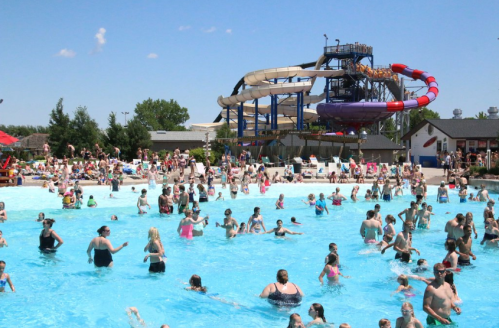 A crowded water park with people swimming in a blue pool and large water slides in the background under a clear sky.