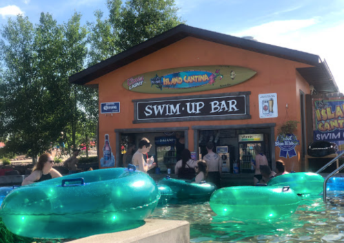 A swim-up bar with people in a pool, surrounded by green inflatable tubes and trees in the background.