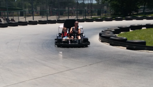 A child and an adult enjoy a go-kart ride on a racetrack, navigating a curve surrounded by tires.
