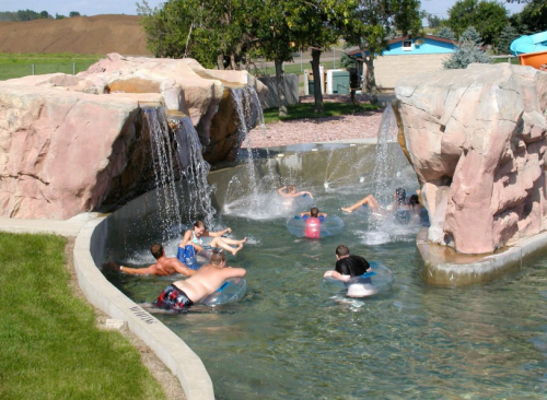 A water park scene with people floating on inner tubes in a pool surrounded by rock formations and waterfalls.