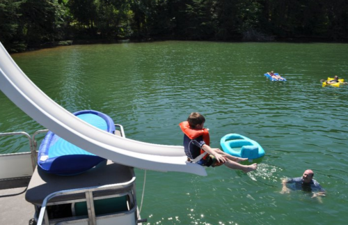 A child prepares to slide into a lake from a boat, while an adult swims nearby and others float on inflatable devices.
