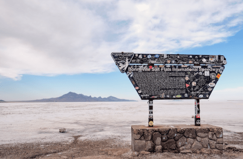 Sign at Bonneville Salt Flats International Speedway, covered in stickers, with a vast salt flat and mountains in the background.