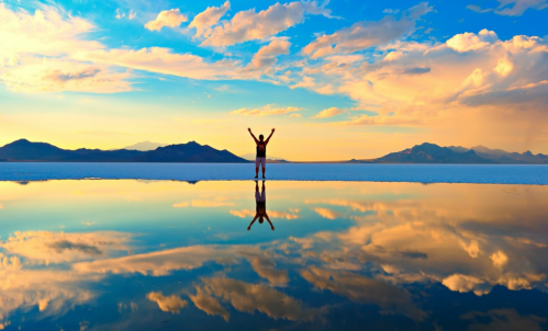 A person stands with arms raised on a reflective salt flat, surrounded by mountains and a colorful sky at sunset.