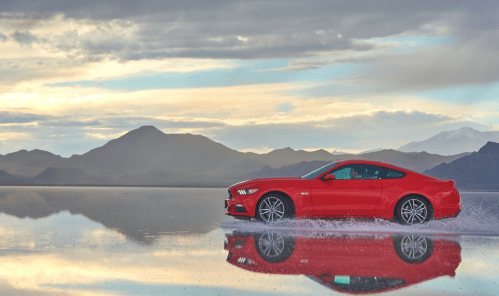 A red sports car splashes through water on a reflective surface, with mountains and a cloudy sky in the background.