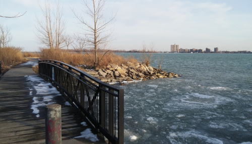A wooden path leads to a rocky shore by a lake, with ice patches and a city skyline in the distance.