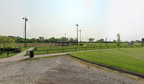 A grassy park with a baseball field, benches, and light poles under a hazy sky.
